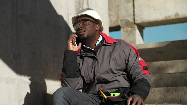 African american worker talks on smartphone. Builder sits on concrete stairs at construction site and speaks on mobile phone, looks around