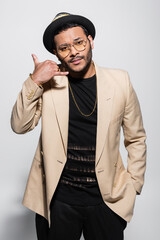 bearded indian hip hop performer in fedora hat and eyeglasses showing call me gesture on grey.