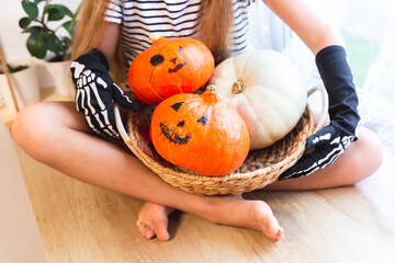 Girl in black gloves with bones sitting cross-legged with basket full of Halloween Pumpkins. Jack-o-lantern. Orange and white pumpkins in basket in hands of child.