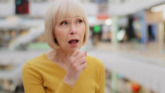 Mature caucasian unwell woman standing indoors coughing covers mouth with hands elderly unhealthy female suffering from flu symptoms colds chronic asthma disease feels choking first symptom of virus