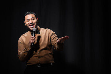 cheerful indian comedian in shirt and bow tie holding microphone during monologue on black. - obrazy, fototapety, plakaty