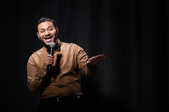 cheerful indian comedian in shirt and bow tie holding microphone during monologue on black.