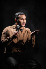 indian comedian sitting and pouting lips while performing stand up comedy into microphone on black.