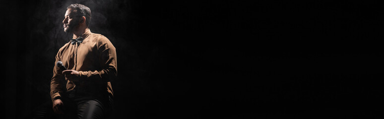 indian comedian holding microphone and looking away on black with smoke, banner.