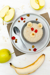 Funny cute kids childrens baby's healthy breakfast lunch oatmeal porridge in bowl look like bear face decorated with apple, banana, dried berry fruits. dessert food art on white wooden table