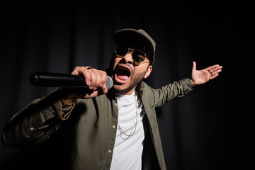 emotional eastern hip hop performer in sunglasses singing in microphone on black.