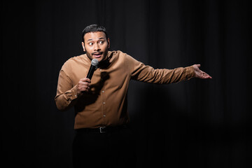 puzzled indian comedian in shirt and bow tie holding microphone and gesturing during monologue on black. - obrazy, fototapety, plakaty