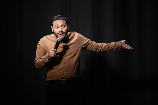puzzled indian comedian in shirt and bow tie holding microphone and gesturing during monologue on black.