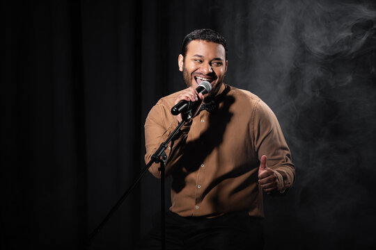 happy indian comedian sitting on chair and performing stand up comedy into microphone on black with smoke.