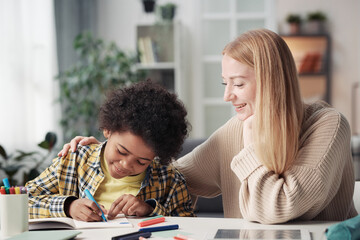 Young mother sitting at table in the room with her adopted son while he drawing with crayons