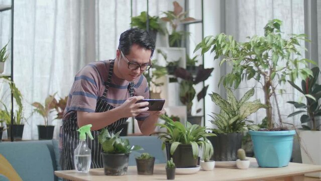 Asian Man Holding Smartphone And Taking Photos Of Plants At Home
