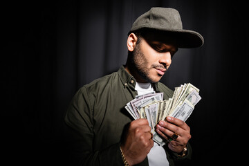 rich indian hip hop performer in cap holding dollar banknotes on black.