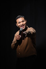 happy indian comedian in shirt and bow tie holding microphone and pointing with finger during monologue on black.