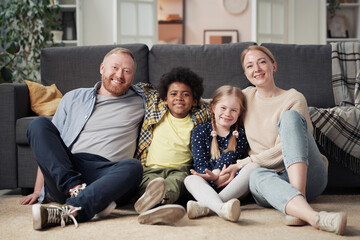 Portrait of happy foster parents sitting on floor with their adopted children and smiling at camera, they resting at home