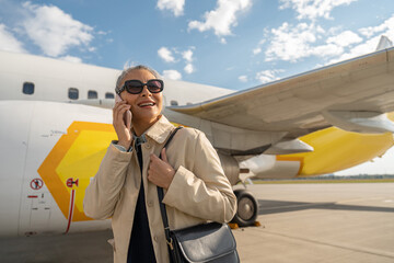 Smiling woman passenger in sunglasses talking phone standing outdoors at airport before boarding