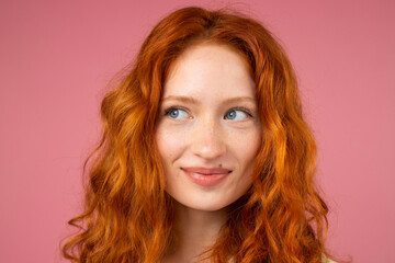 Close up photo of a beautiful redhead lady with blue attractive eyes looking aside over a pink background with smiling facial expression.