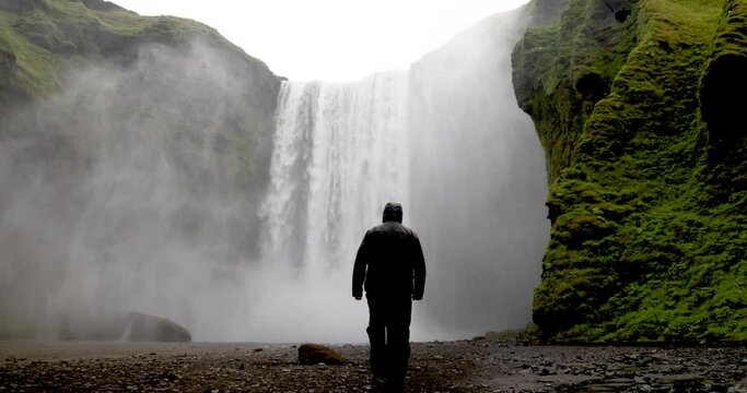 Skogafoss waterfalls in Iceland with man in rain jacket walking towards falls in slow motion.