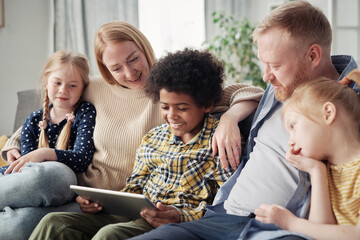 Happy adopted children watching video on digital tablet together with their foster parents during leisure time on sofa at home