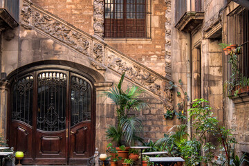 Ornaments of a medieval inner yard in the Gothic Quarter of Barcelona, Catalonia, Spain, Europe. Historical stone stairs, walls and beautiful ancient patterns. Plants, flower pots, an old wooden door.