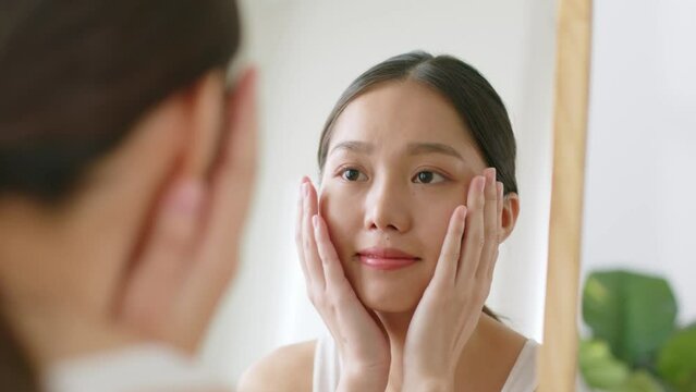 Young Asian woman touching healthy facial skin look at mirror
