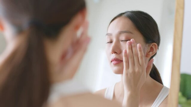 Young Asian woman apply serum and moisturizing cream on healthy facial skin and look at mirror