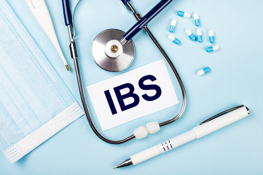 On a blue background, a stethoscope, a medical mask, pills, a pen and a white card with the text IBS Irritable Bowel Syndrome. View from above.