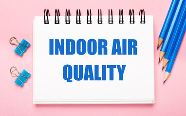 On a light pink background, light blue pencils, paper clips and a white notebook with the text INDOOR AIR QUALITY