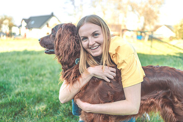 Woman happily smiling at playing with her red Irish setter dog outdoors on fresh green grass in the...