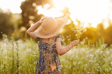 smiling child girl in a big mommys straw hat with bouquet of wildflowers in a green grassy meadow on summer sunny day. Happy childhood concept. Copy space.