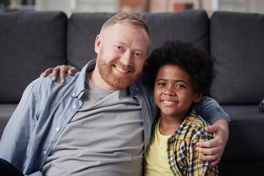 Portrait of dad and adopted son embracing and smiling at camera sitting at home