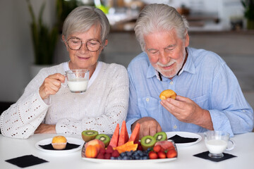 Beautiful caucasian retired senior couple having breakfast at home with fresh seasonal fruit, milk and cupcake, healthy eating concept