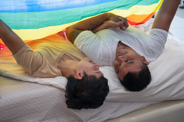 Happy Asian homosexual gay couple with rainbow flag covering and lying on bed together..LGBTQ,...