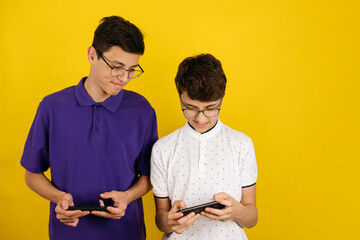 Two cute young men in eyeglasses with phones in their hands look at each other's screen, yellow background