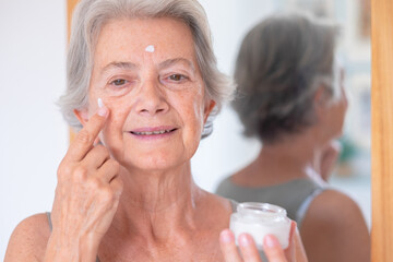 Close up shot of senior beautiful woman applies anti aging cream on wrinkled face - elderly lady...