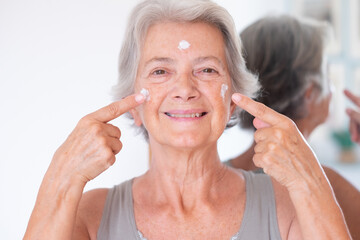 Portrait of happy senior beautiful woman applies anti aging cream on wrinkled face - elderly...