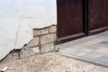 Bricks are visible in the old wall of the building next to the metal door, due to peeling off plaster. A concrete path leads to the entrance to the house.