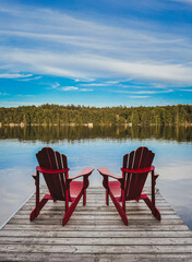 Two red adirondack chairs on the end of a dock overlooking a lake.