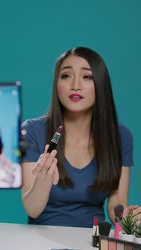 Vertical video: Beauty blogger recording product recommendation on camera, filming lipstick review with makeup cosmetics in studio. Female influencer broadcasting video tutorial for online social