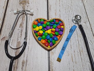 Stethoscope, measuring tape and a heart-shaped bowl with colorful candies on a wooden table.