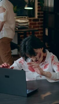 Vertical video: Brain dead spooky zombie working on modern computer. Brain-eating creppy dead walking corpse with bloody and deep wounds sitting at desk in office while trying to use laptop for