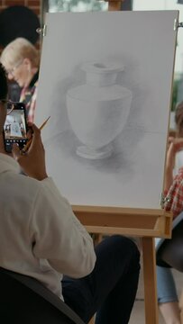 Vertical video: Creative student taking picture of vase sketch on smartphone in art class. Young man using mobile phone to take photo and remember drawing skills technique at educational lesson.