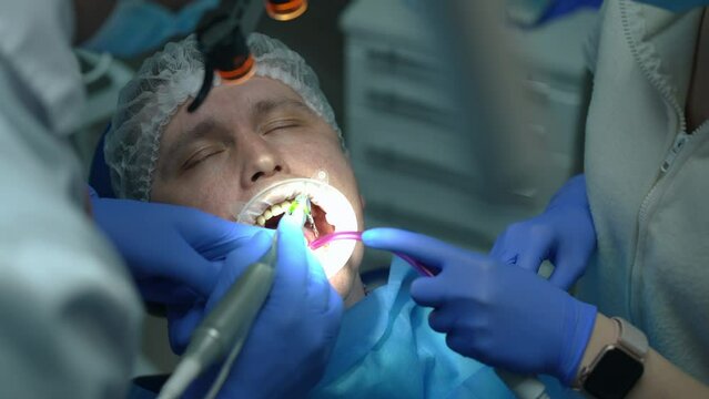 Close-up portrait of Caucasian patient in dental chair closing eyes as doctor drilling ill tooth and assistant using saliva ejector in slow motion. Man undergoing treatment in hospital indoors