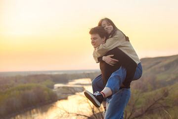 a beautiful young girl sat on the back of a guy, they run and have fun, relaxing outside the city on a walk in the rays of the setting sun. Love and positive emotions in young people