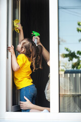 mother teaches her little daughter to clean up the house, they wash the window in their house together with a cleaning agent and a yellow rag for cleaning the house