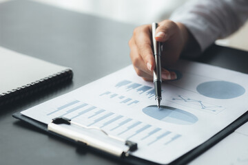 Close-up businesswoman's hand holding a pen pointing at a bar chart on a corporate financial information sheet, the businesswoman examines the financial information provided by the finance department.