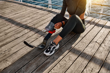 Cropped photo of woman with prosthetic leg sitting after sports training outdoors and adjusting prosthesis. A concert of strength, sport and motivation.