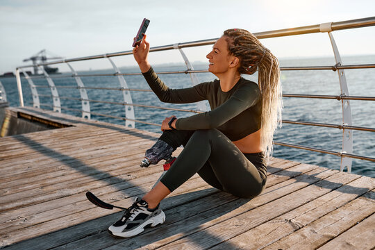 A woman in sportswear with a prosthetic leg sitting after sports training in the fresh air and taking a photo of herself. A concert of strength, sport and motivation.