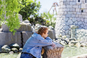 A woman sitting under a willow looks at the river near the stone castle in the amusement park. Spring tranquility. Park decorations. Selective focus.