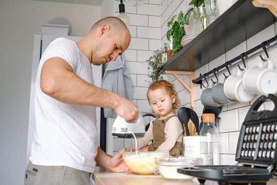 A dad and baby toddler cooking together. A father and kid have fun spending time together on the kitchen baking waffles at home. A man mixing ingredients for dough using blender.