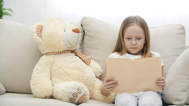 4k video of small girl sitting with teddy bear and holding a book.